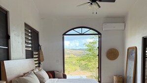 A bedroom with a bed, a ceiling fan, a window with a view of the outdoors, and a wall-mounted air conditioner. - Largest Private Estate on Vieques — 30 Acres, 3 Homes, the Island's Largest Pool (Vieques)