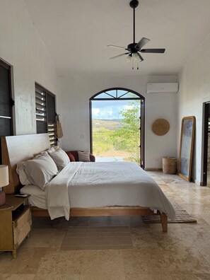 A bedroom with a bed, a ceiling fan, a window with a view of the outdoors, and a wall-mounted air conditioner. - Largest Private Estate on Vieques — 30 Acres, 3 Homes, the Island's Largest Pool (Vieques)