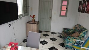 Et lille værelse med et fjernsyn ophængt på væggen, et træsidebord med en potteplante, en stribet sofa og en stol med en blomstret pude.