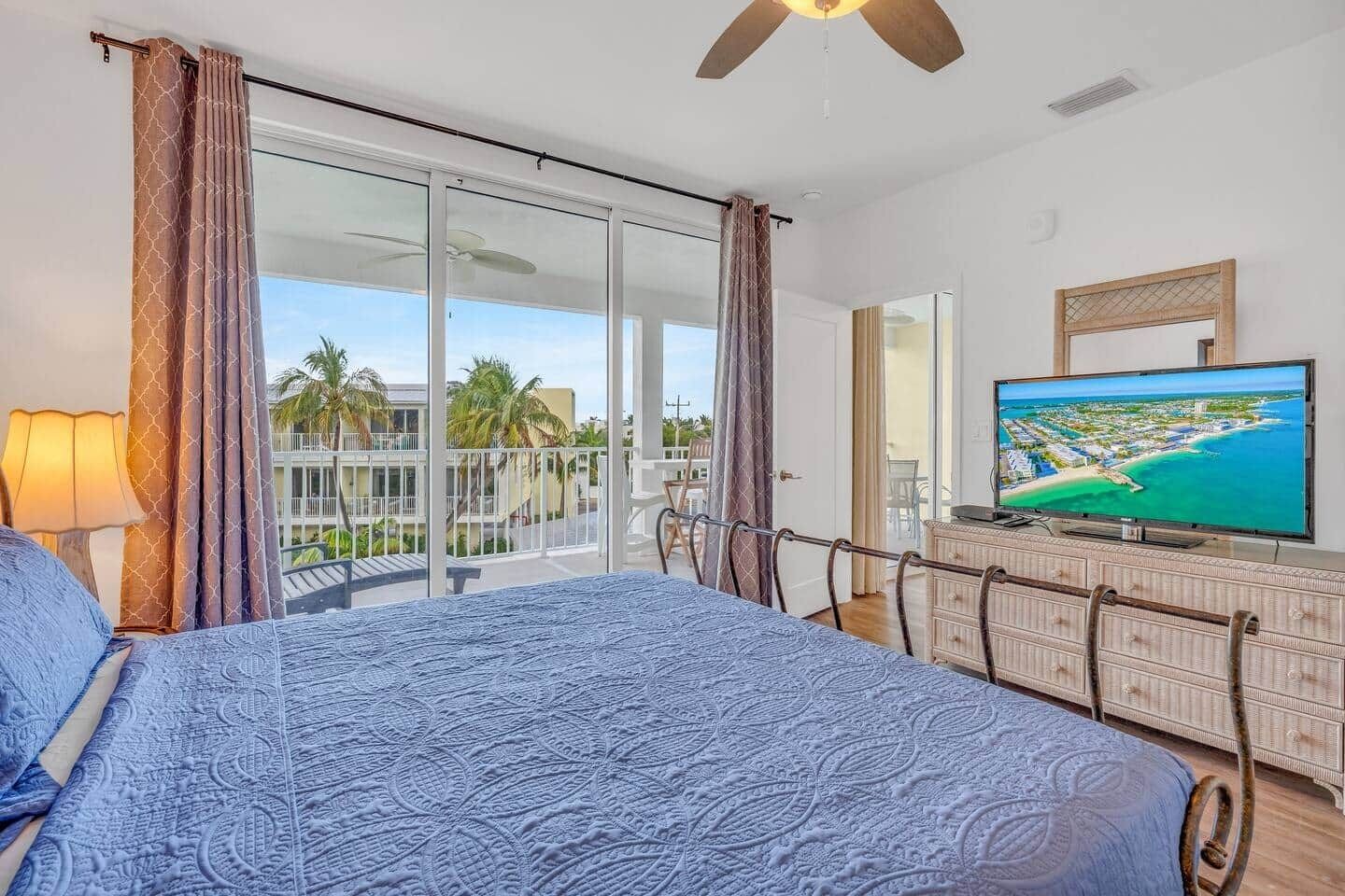A bedroom with a bed, a television, a dresser, and a balcony with a view of a beach and palm trees.