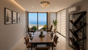 A modern dining area with a large table, chairs, a chandelier, a potted plant, and a view of the sea.
