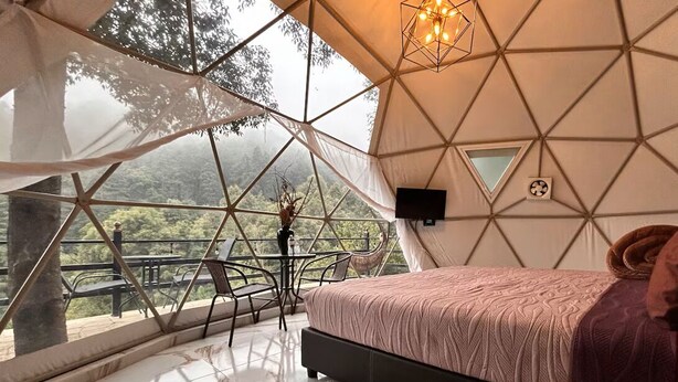 A bedroom with a bed, a TV, a table, and a chair, with a view of trees through a geometric window. - FEYRE · Meraki Cabañas & Glamping by GEstores (Mineral del Chico)