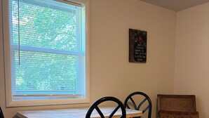 Zona pranzo con tavolo, quattro sedie, finestra con persiane, quadro appeso al muro e una sedia in vimini.