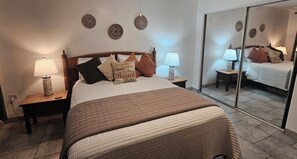 A bedroom with a bed, two bedside tables with lamps, a mirror, and decorative wall art. - Lovely 1-bedroom apartment in Napili, perfect for your beach getaway (Lahaina)