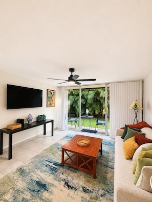 A modern living room with a flat-screen TV, a ceiling fan, a wooden coffee table, a sofa with colorful cushions, and a view of a garden through large sliding doors.