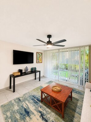 A living room with a flat-screen TV, a ceiling fan, a coffee table, and a view through a sliding glass door.