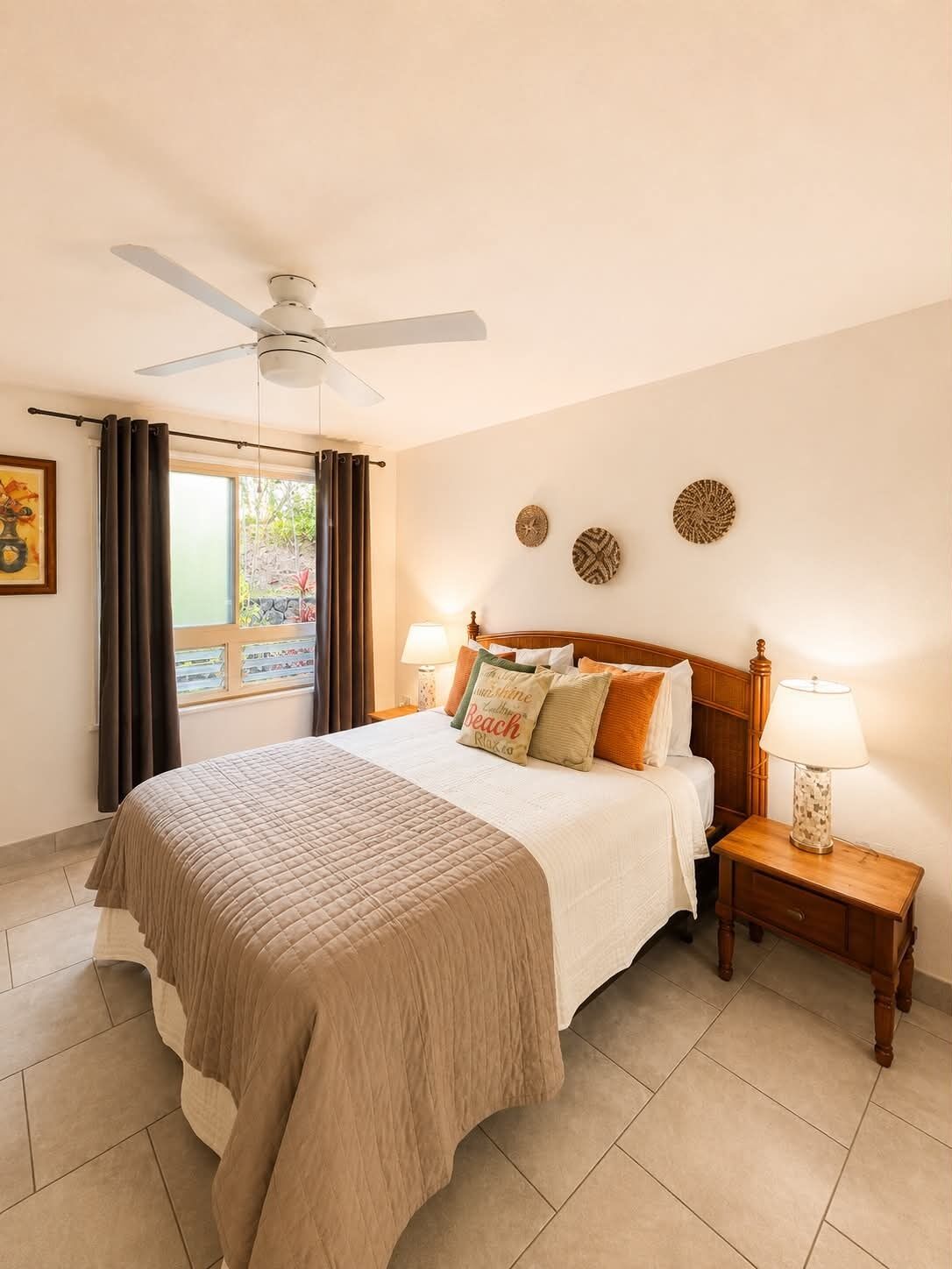 A hotel room with a bed, a ceiling fan, a window with curtains, two bedside tables with lamps, and wall decor.