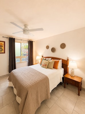 A hotel room with a bed, a ceiling fan, a window with curtains, two bedside tables with lamps, and wall decor.