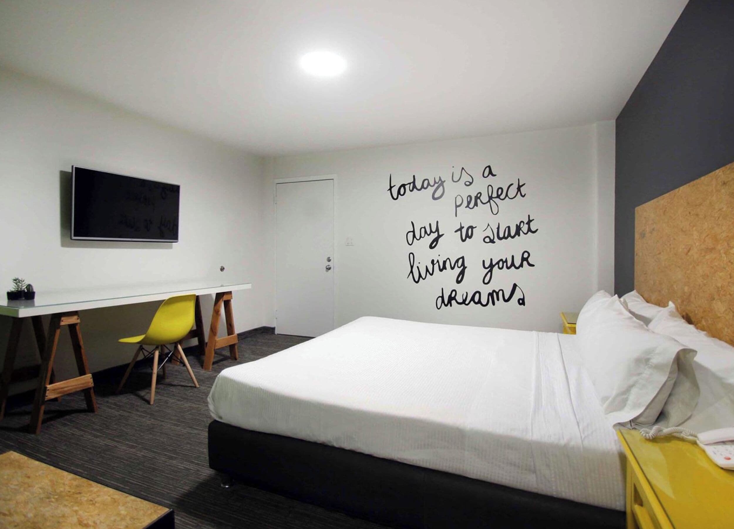 Habitación de hotel con cama, escritorio, silla, televisor y una cita motivacional en la pared.