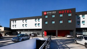 Hotel Stay Río, con un diseño moderno, presenta un letrero rojo y negro llamativo, amplias ventanas y un estacionamiento con varios automóviles.