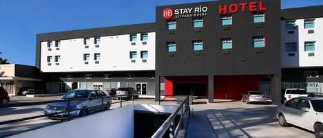 Hotel Stay Río, con un diseño moderno, presenta un letrero rojo y negro llamativo, amplias ventanas y un estacionamiento con varios automóviles.