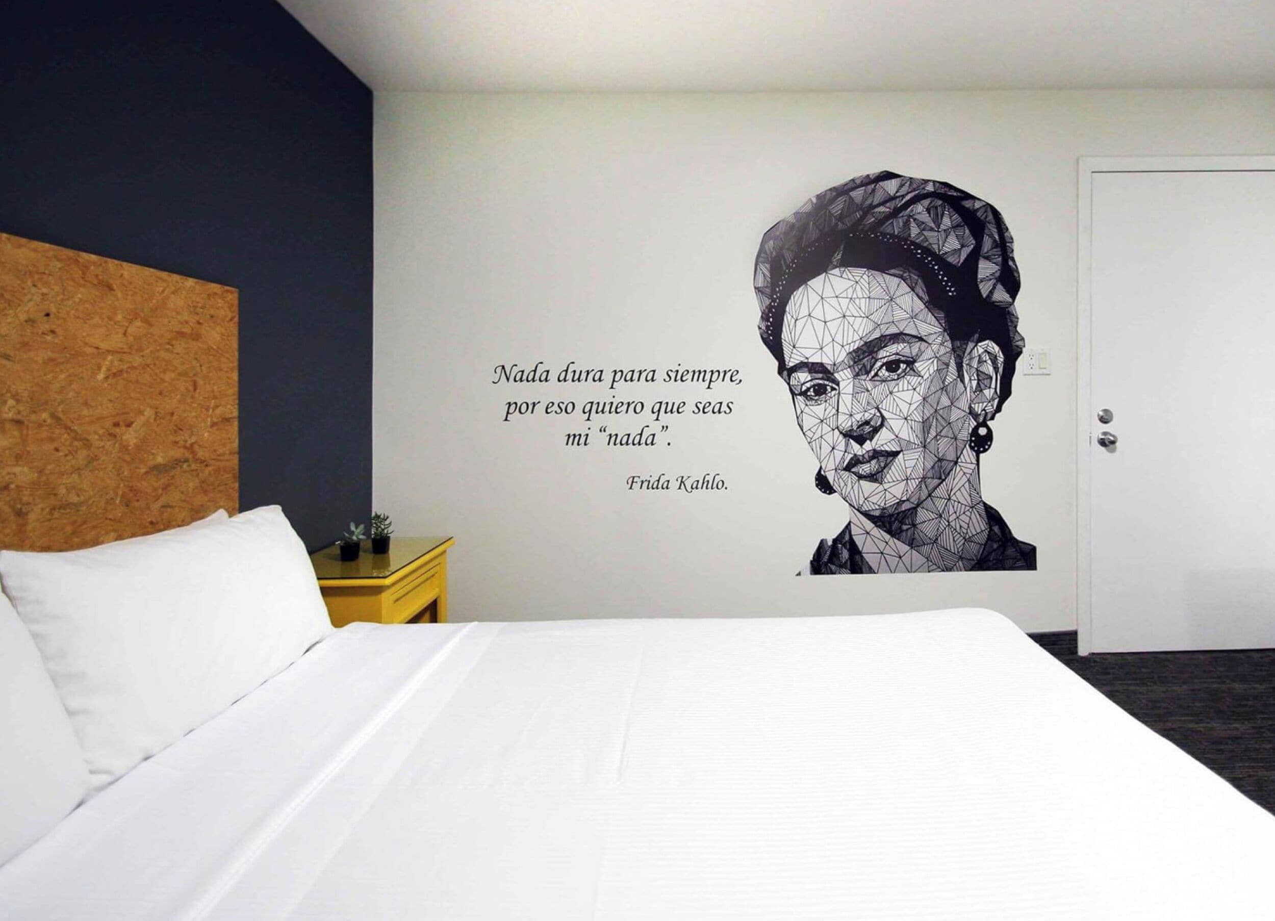 A hotel room with a bed, a wall with a quote, a wooden headboard, a nightstand, and a door.