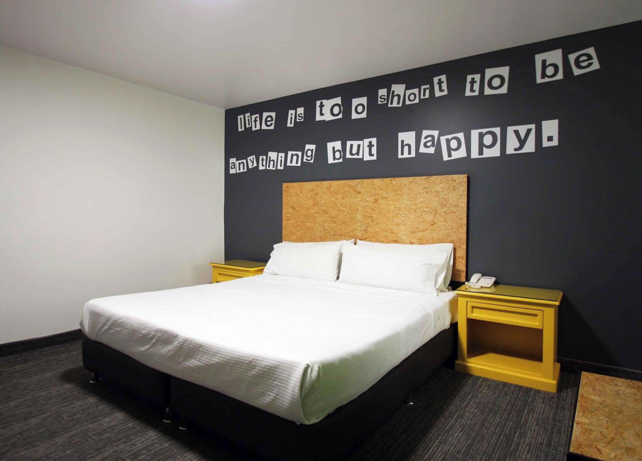 A hotel room with a bed, a headboard, two bedside tables, and a wall with a motivational quote.