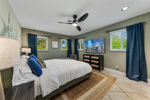 Une chambre à coucher avec un lit, un ventilateur de plafond, une télévision, un bureau et une fenêtre avec des rideaux.