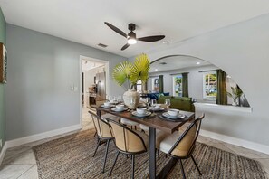 Un coin repas avec une table dressée pour quatre personnes, un ventilateur de plafond et une vue sur la cuisine et le salon, séparés par une arche.