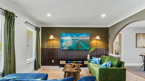 Ein Wohnzimmer mit einem grünen Sofa, blauen Kissen, einem Couchtisch und einem großen Fernseher an der Wand.