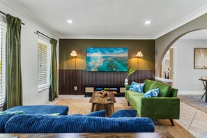 Ein Wohnzimmer mit einem grünen Sofa, blauen Kissen, einem Couchtisch und einem großen Fernseher an der Wand.