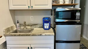 Cozinha com armários brancos, geladeira de aço inoxidável, micro-ondas, cafeteira e uma prateleira com objetos decorativos.
