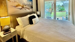 Quarto com cama, mesa de cabeceira, luminária, quadro e vista para o exterior.