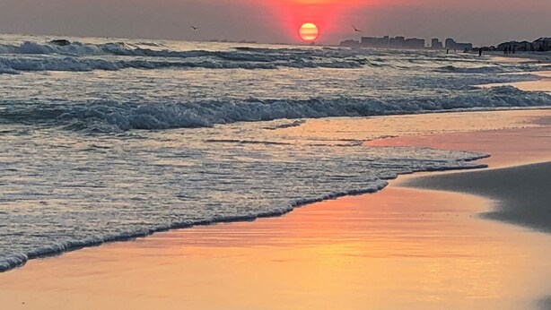 Uma praia com ondas, um pôr do sol e o horizonte distante de uma cidade.