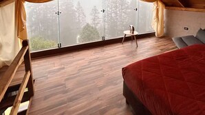 A bedroom with a sloped ceiling, a bed, a small table with a chair, and a large window with curtains. - INARI · Meraki Cabañas & Glamping by GEstores (Mineral del Chico)