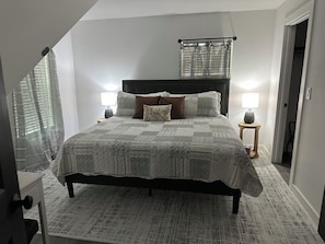 Un dormitorio con una cama, dos mesitas de noche, un ventanal con persianas y una puerta.