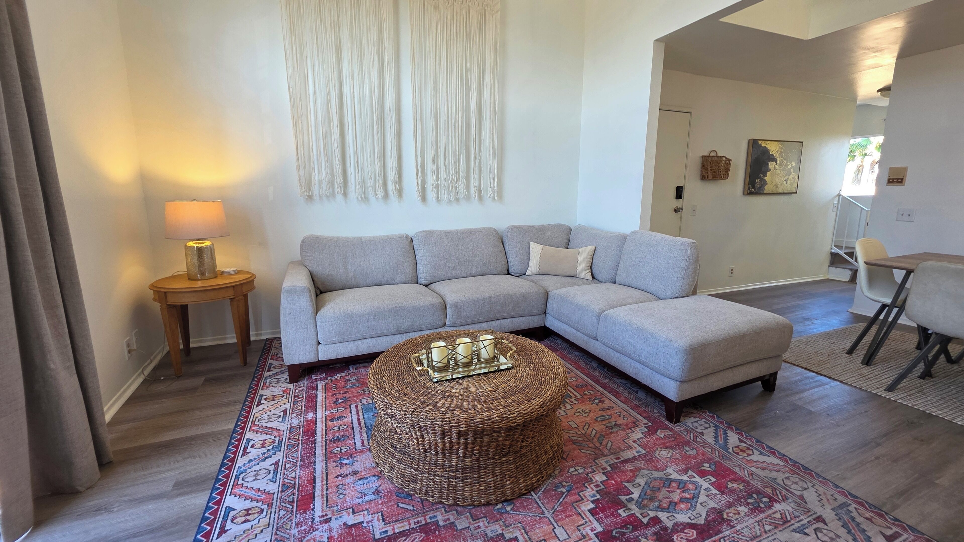 A living room with a grey sectional sofa, a round wicker ottoman, a patterned rug, and a small side table with a lamp.