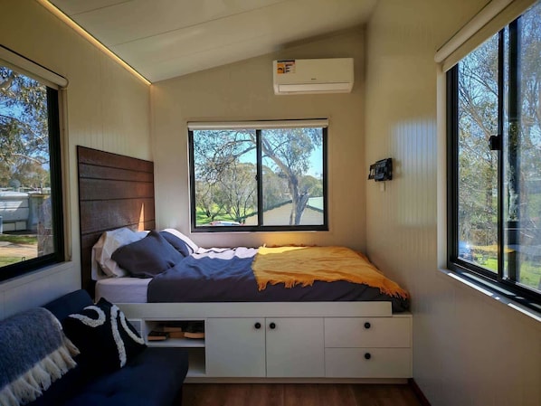 A bedroom with a bed, a window, a built-in storage unit, and an air conditioning unit. - The Frosty Trout - Lakeside Snowy Mts Tiny House (Braemar Bay)