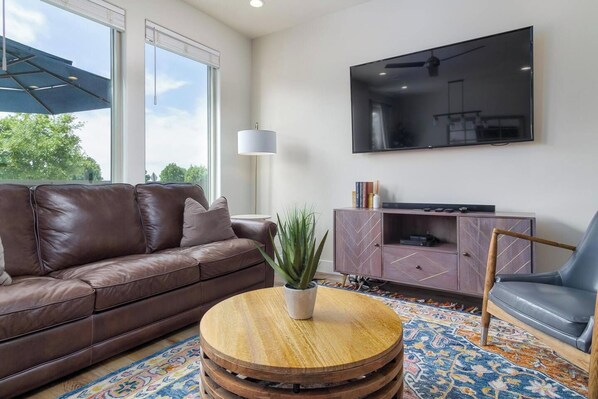 A modern living room with a brown leather sofa, a round wooden coffee table with a plant, a grey leather armchair, a TV mounted on the wall, and a patterned rug.