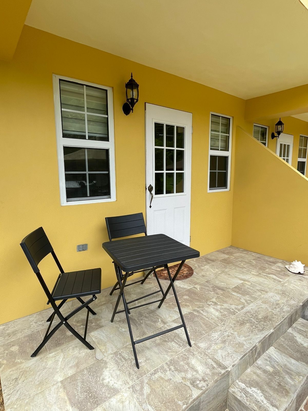 Paredes externas amarelas com uma porta e janelas brancas, uma mesa dobrável preta com uma cadeira e um tapete pequeno em um pátio com azulejos.