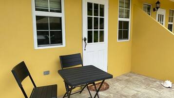 Paredes externas amarelas com uma porta e janelas brancas, uma mesa dobrável preta com uma cadeira e um tapete pequeno em um pátio com azulejos.