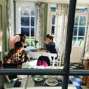 Un coin repas avec une table dressée pour deux personnes, une fenêtre avec des rideaux et une vue sur la verdure à l’extérieur.