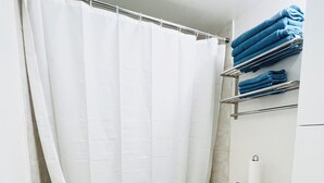 A bathroom with a white shower curtain, a white toilet, a blue trash bin, and a shelf with folded towels. - Cozy Vibes Condo (Toronto)