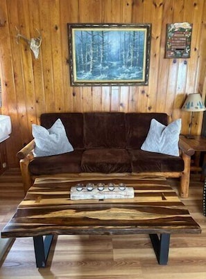 Interior em madeira com um sofá marrom, uma mesa de centro de madeira e um quadro na parede.