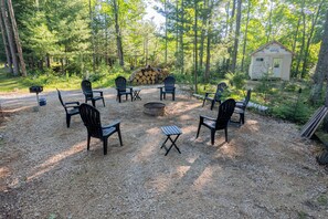 Ein rustikaler Außenbereich zum Sitzen mit Stühlen und einem Feuerkorb, umgeben von einem Wald.