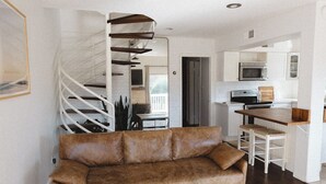 Una sala de estar moderna con un sofá de cuero marrón, un tapete blanco y una escalera espiral que sube a un nivel superior.