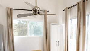 Una habitación con una mesa redonda, cuatro sillas, un ventilador de techo, un armario y una ventana con cortinas.