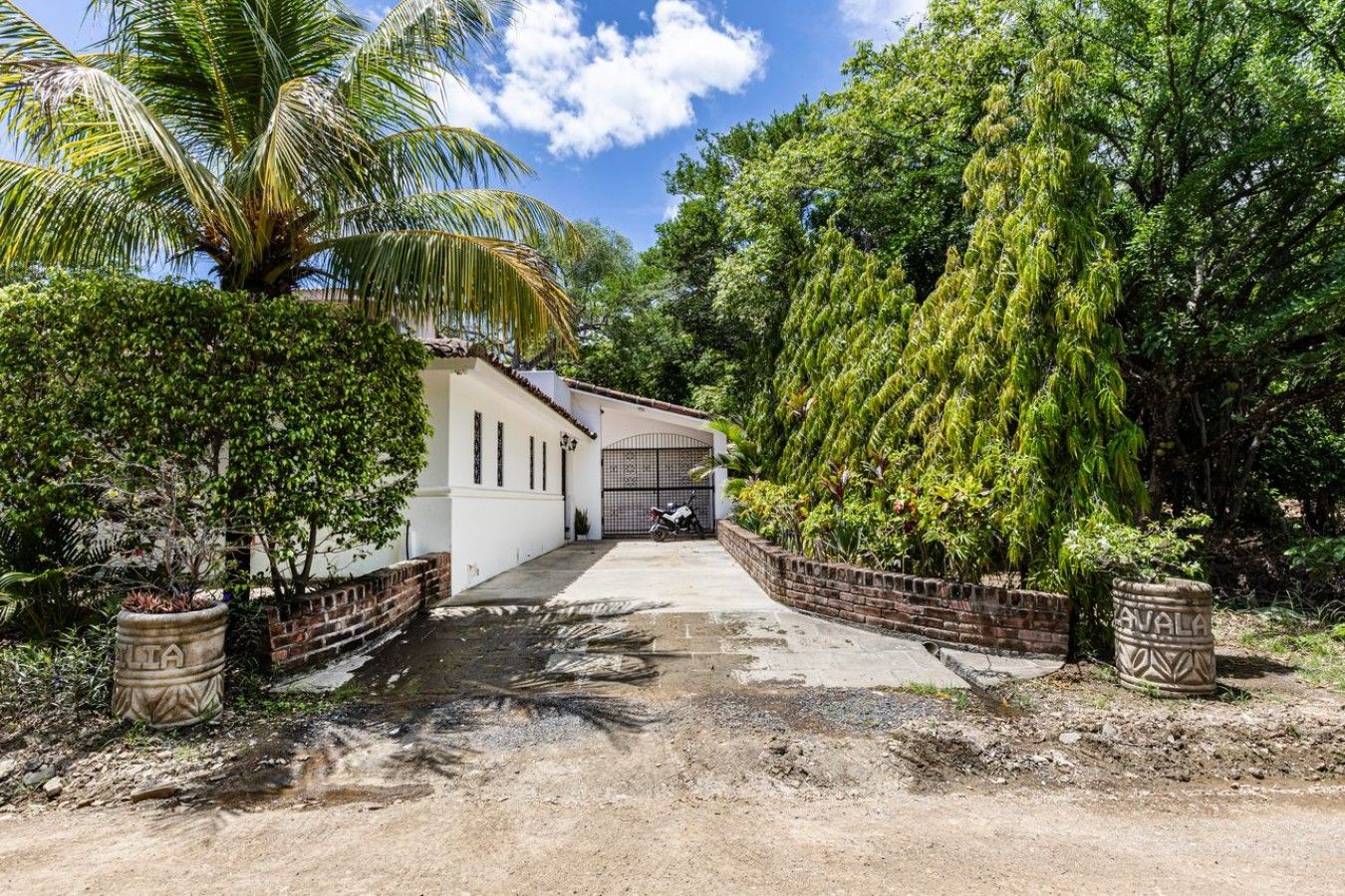 En stig leder fram till en vit byggnad med en grind som ingång, omgiven av frodig grönska och palmer.