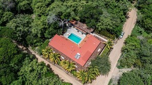 An aerial view of a property with a swimming pool, a red-roofed building, and surrounding dense greenery.
