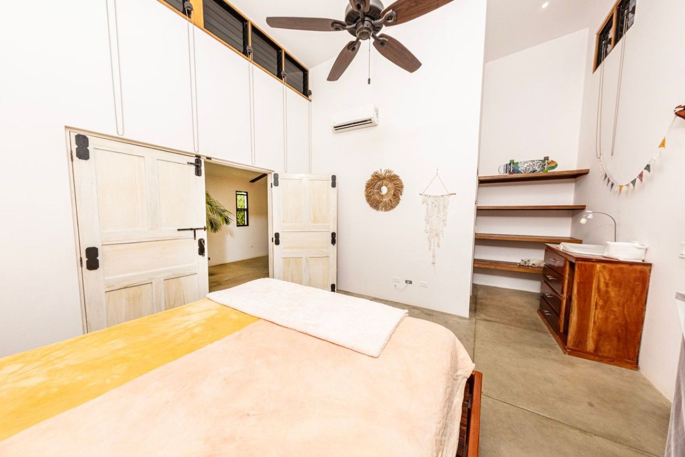 A bedroom with a bed, a ceiling fan, a wardrobe, a desk, and a small bathroom area.