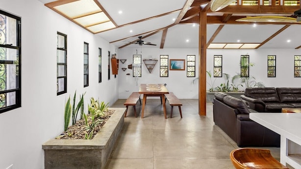 Un intérieur spacieux avec de hauts plafonds, des poutres apparentes, un coin repas, un canapé et un ventilateur de plafond.