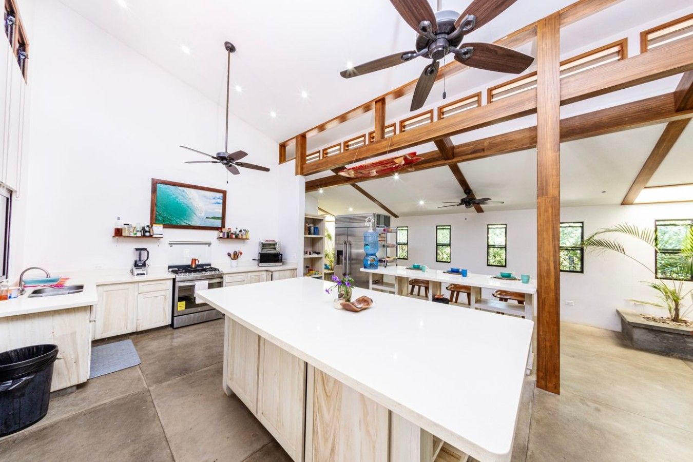 Una cocina amplia con isla central, vigas de madera, ventiladores de techo y un ventanal grande.