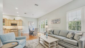 A spacious living area with a sofa, a coffee table, and a dining table. There is a kitchen area with cabinets and a microwave. The room has a large window with blinds and a sign that reads 'Anna Maria Island'. - Hidden Palms - Private Pool, Quiet Island Retreat Near Beach (Holmes Beach)