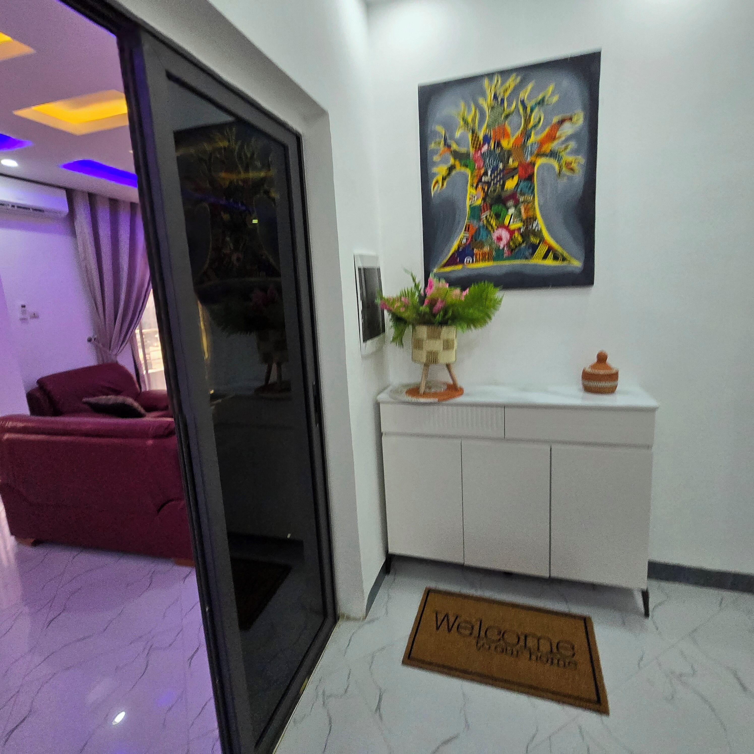 A modern hotel lobby with a glass door entrance, a sofa, a white console table with a vase of flowers, and a colorful abstract painting on the wall.