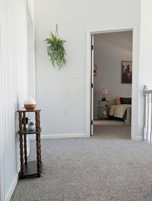 Ein Flur mit Pflanze, einem kleinen Tisch mit Vase und einer Tür zu einem Schlafzimmer mit Bett und Nachttisch.