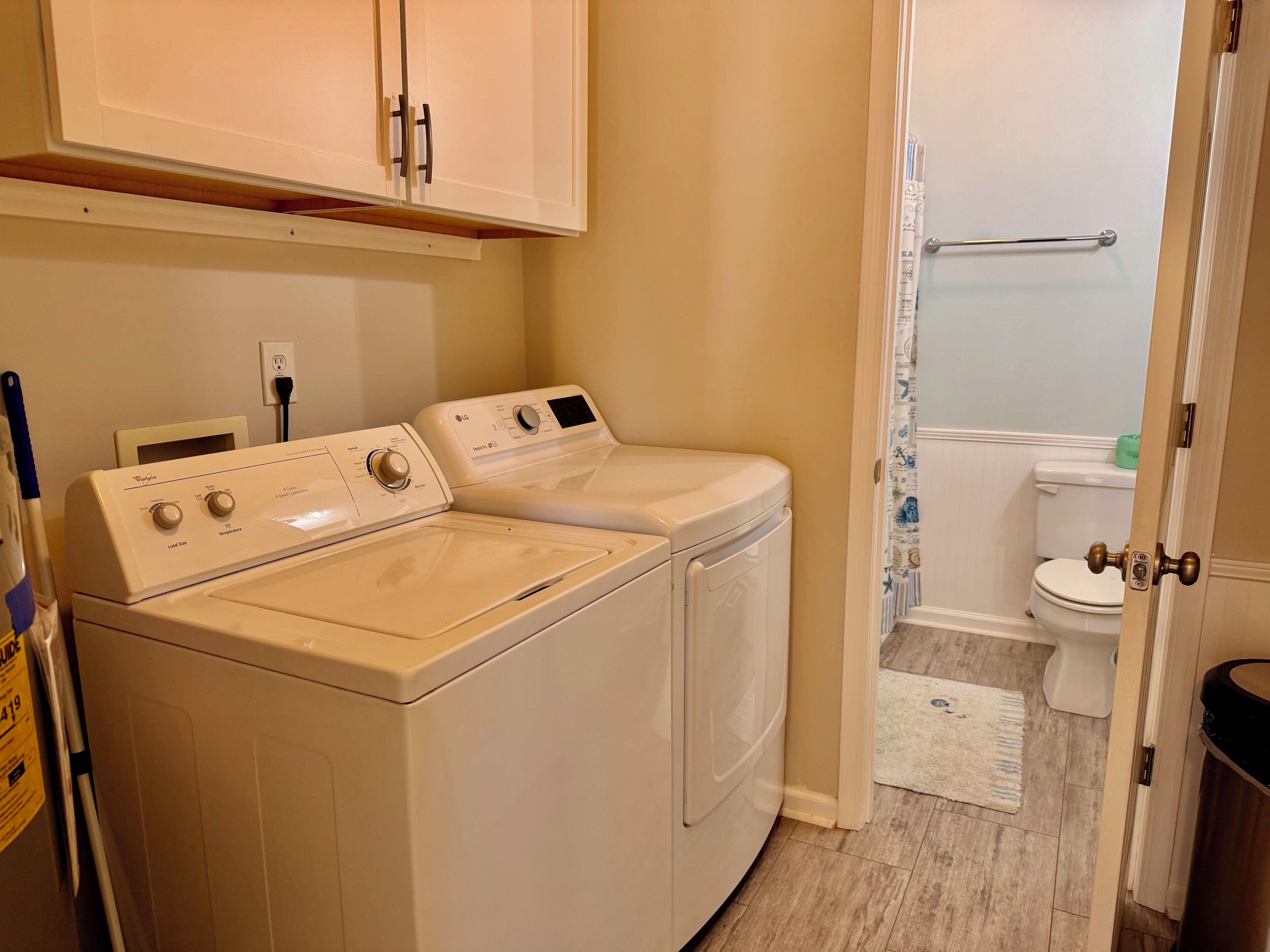 Een wasserette met twee voorladers en drogers, een kast erboven, en een deur naar een badkamer met toilet en douchegordijn.