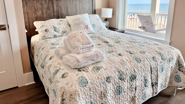 A bedroom with a bed, pillows, a nightstand, a lamp, and a view of the ocean through a window. - Oceanfront 8BR Oak Island home with private heated pool. (Oak Island)
