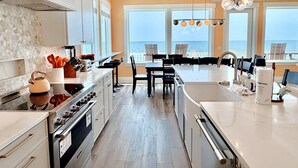 A modern kitchen with a view of the sea, featuring stainless steel appliances, a dining area with chairs, and a large island with a sink. - Oceanfront 8BR Oak Island home with private heated pool. (Oak Island)