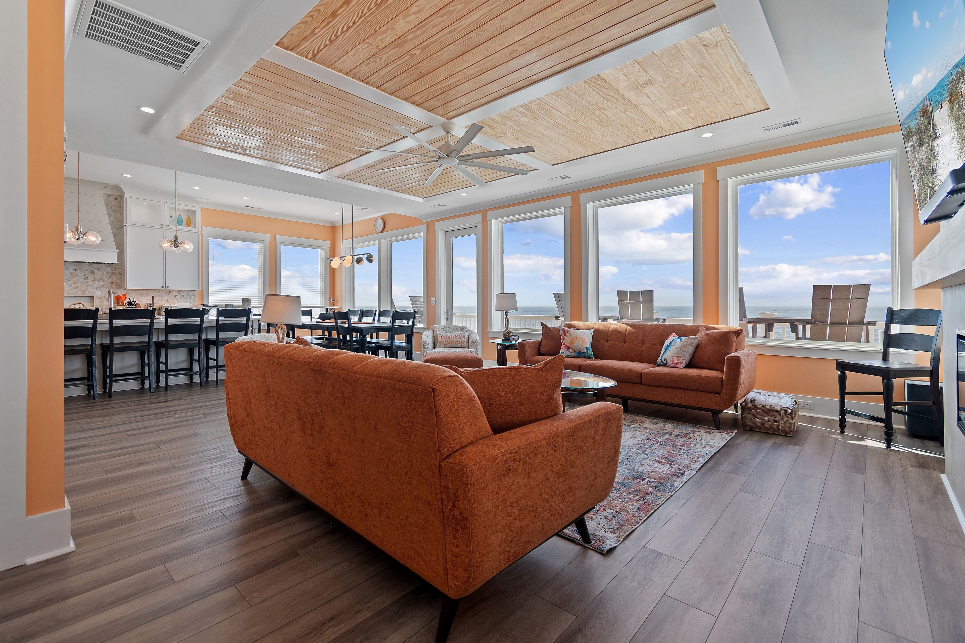 A spacious living area with a large window overlooking the beach, a ceiling fan, and a dining area with a view.