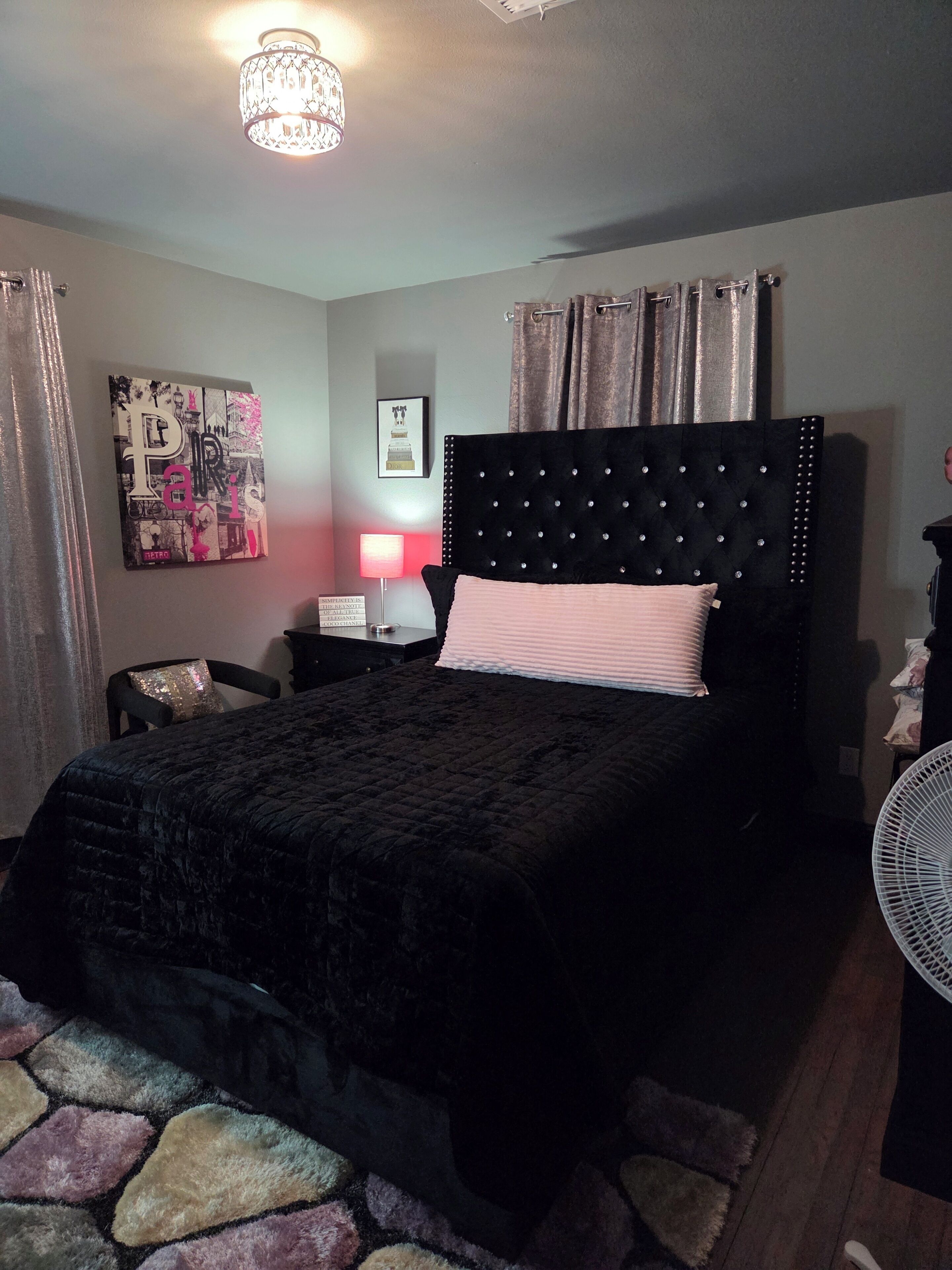 Une chambre à coucher avec une tête de lit capitonnée, un lit avec un oreiller rose, une table de chevet avec une lampe, une horloge murale et une affiche.
